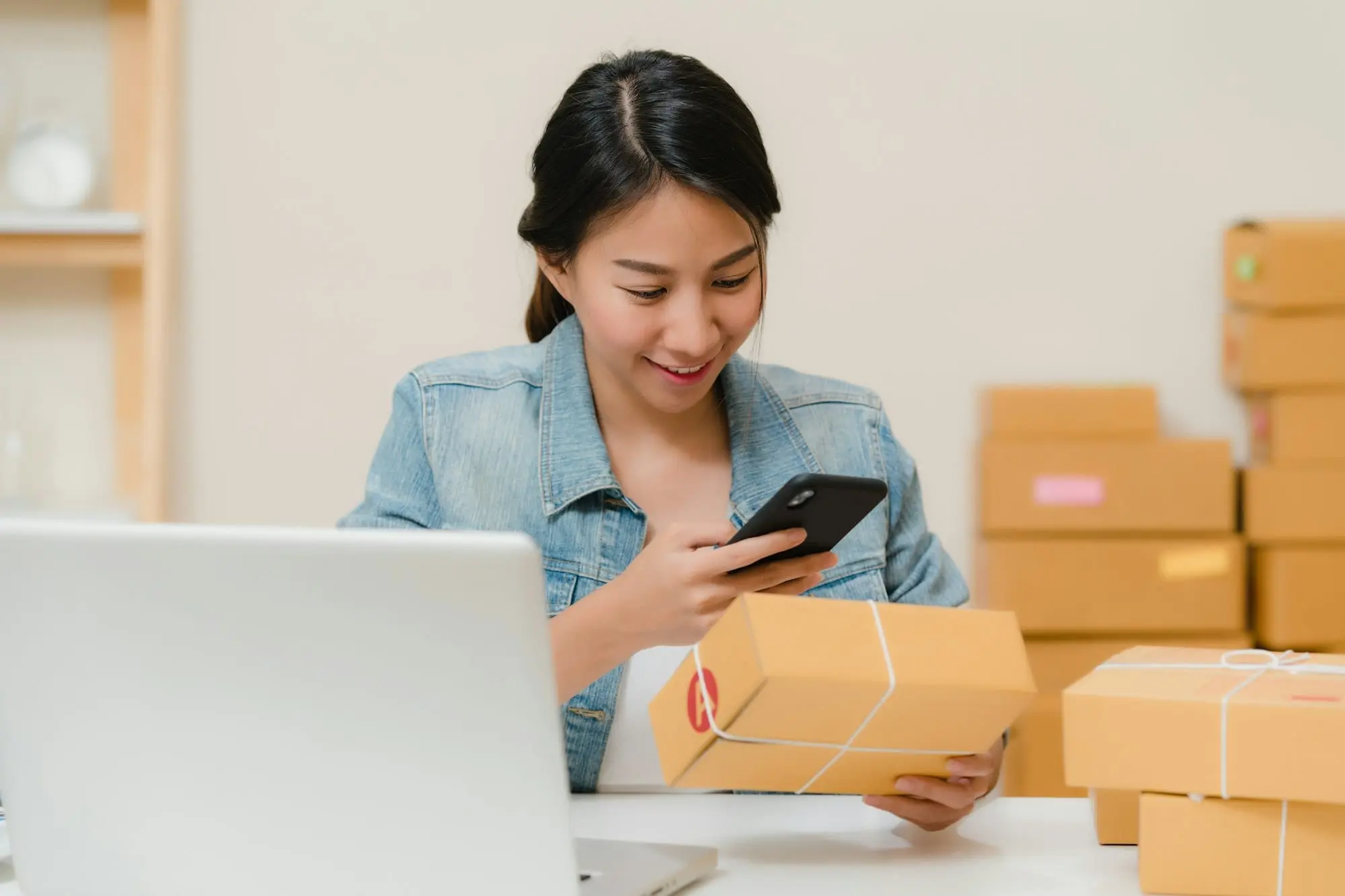 A small business owner checking a parcel against an order on a phone beside a laptop and stacked boxes